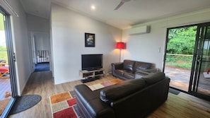 Ein Wohnzimmer mit einer Couch, einem Fernseher und einer roten Lampe. Eine Tür führt in einen anderen Raum, und es gibt einen Blick ins Grüne nach draußen.