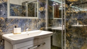 Bagno con parete in marmo, cabina doccia in vetro e lavandino bianco con dispenser per sapone.