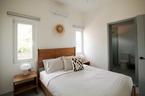Une chambre avec un lit en bois, des tables de chevet, un climatiseur fixé au mur et une vue sur la salle de bain par une porte ouverte.