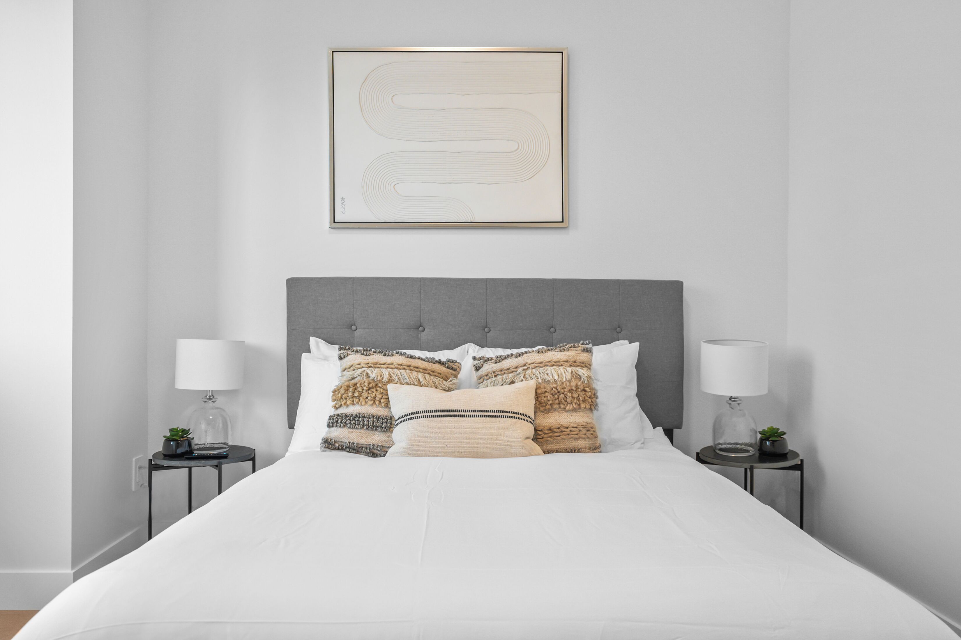 Una cama bien hecha con cabecera gris, dos mesitas de noche con lámparas y una obra de arte abstracta enmarcada sobre la cama.