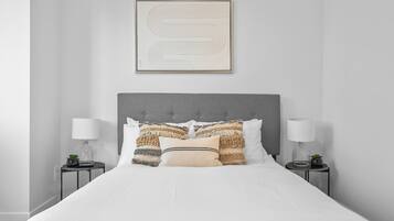 Una cama bien hecha con cabecera gris, dos mesitas de noche con lámparas y una obra de arte abstracta enmarcada sobre la cama.
