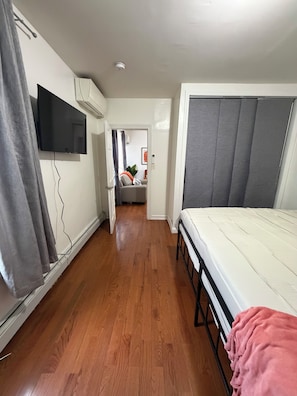 A narrow hallway with a bed on the right, a TV on the left, and a door leading to another room.