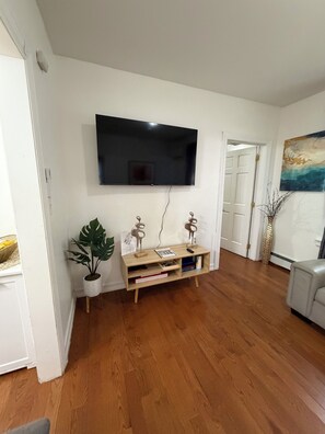 A living room with a flat-screen TV mounted on the wall, wooden flooring, a potted plant, a wooden entertainment unit with books and decorative items, and a painting on the wall.