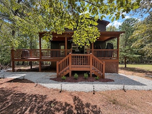 A wooden cabin with a deck, surrounded by trees and gravel.