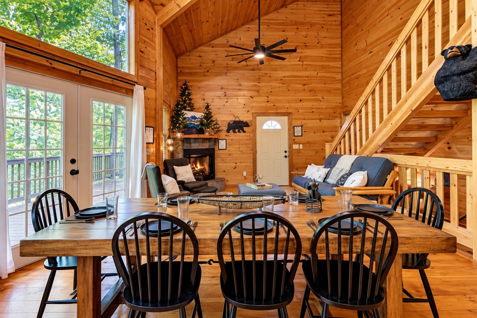 A cozy cabin interior with a dining area, a fireplace, a staircase, and a view of the outdoors.