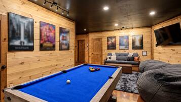 A pool table in a room with wooden walls, a sofa, and a television mounted on the wall.