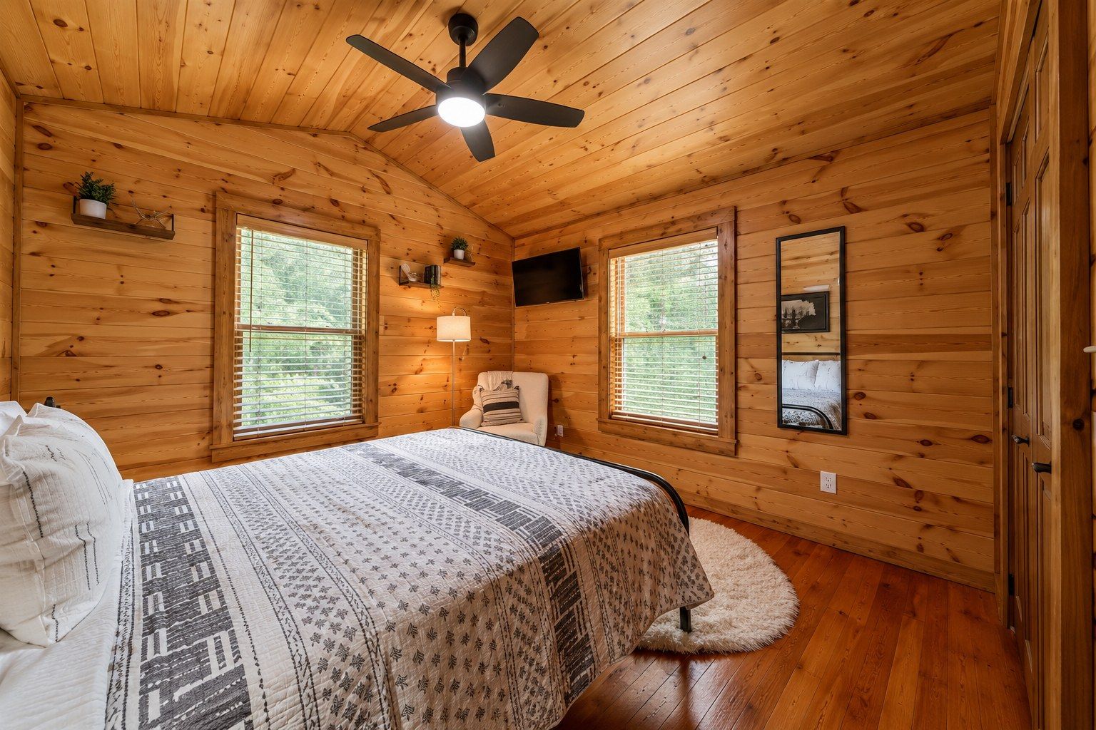 A wooden cabin bedroom with a bed, a chair, a ceiling fan, and two windows.