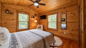 A wooden cabin bedroom with a bed, a chair, a ceiling fan, and two windows.