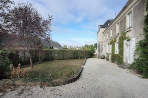 Une allée de gravier mène à un bâtiment à la façade traditionnelle, entouré d’une haie et d’arbres bien entretenus.