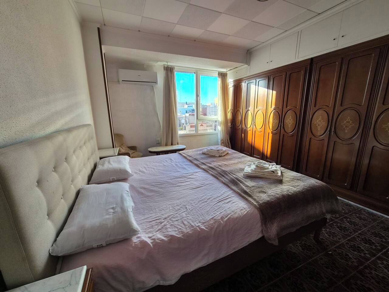 Habitación de hotel con una cama grande, un armario de madera, una ventana con cortinas y vistas a la ciudad.