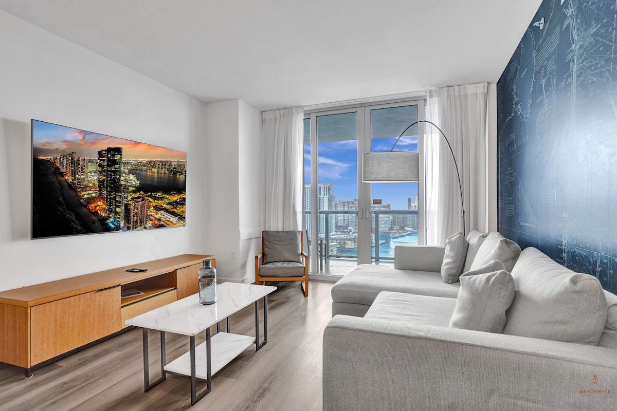 A modern living room with a large flat-screen TV, a sofa, a coffee table, and a view of a cityscape through large windows.