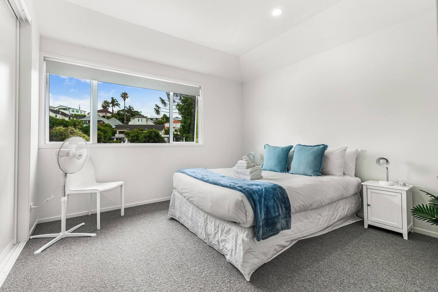 A bedroom with a bed, a bedside table, a chair, a standing fan, and a window with a view of houses and trees.