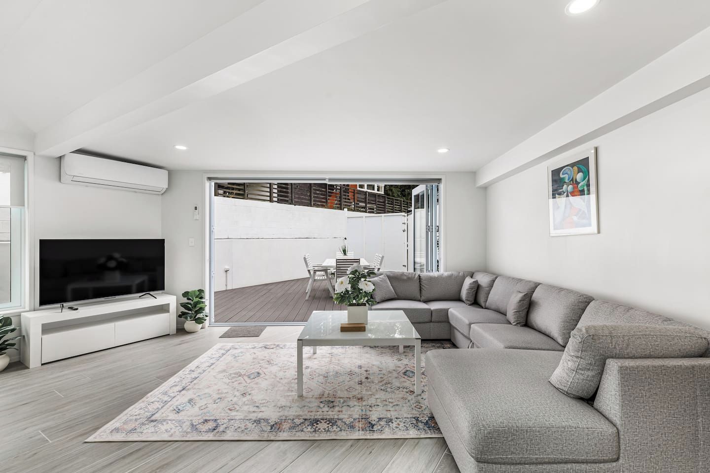 A modern living room with a grey sofa, a flat-screen TV, and a dining area visible through a glass door.