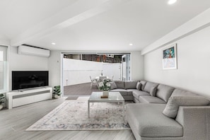 Een moderne woonkamer met een grijze bank, een flatscreen televisie en een eethoek zichtbaar door een glazen deur.