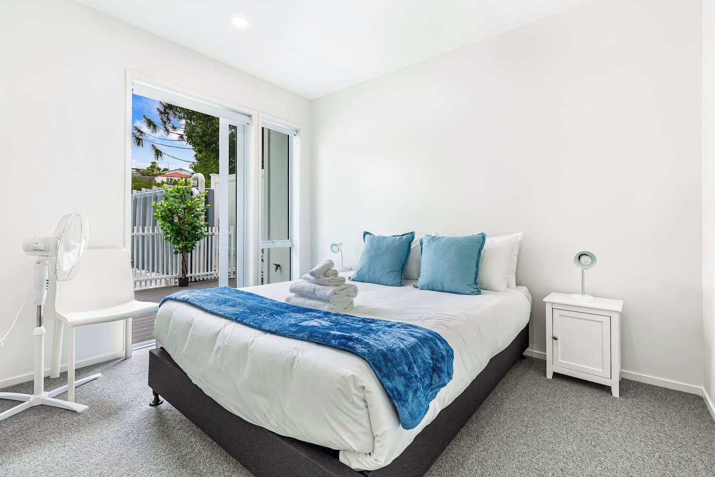 A modern bedroom with a bed, bedside tables, a fan, and a view of the outside.