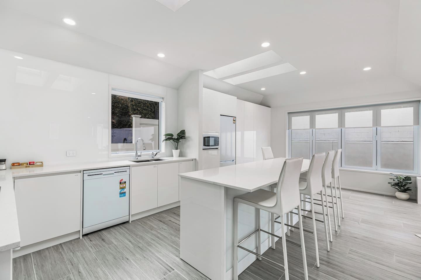 A modern kitchen with white cabinetry, a built-in dishwasher, a dining area with white chairs, and a large window providing natural light.