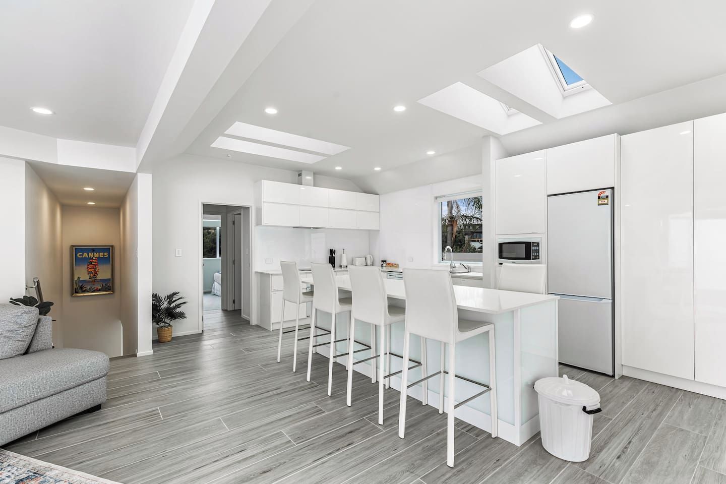 A modern kitchen with white cabinetry, a dining area with white chairs, and a grey sofa in a living room.