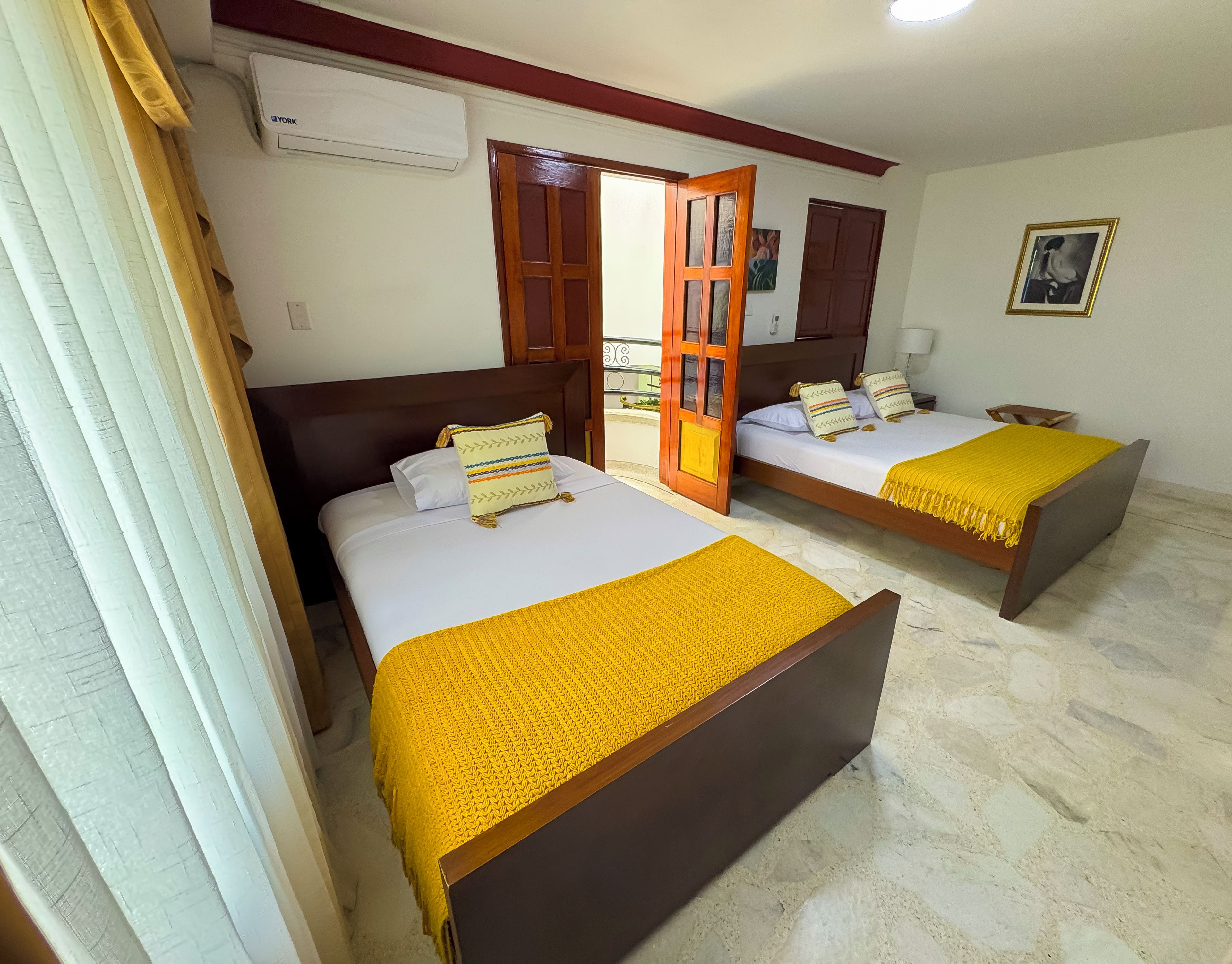 Quarto de hotel com duas camas, cabeceira de madeira, colcha amarela e janela com cortinas.