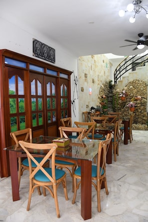 Un espace repas avec des chaises et des tables en bois, un mur décoratif orné d’une image de Bouddha et un mur en pierre agrémenté de plantes en pot.