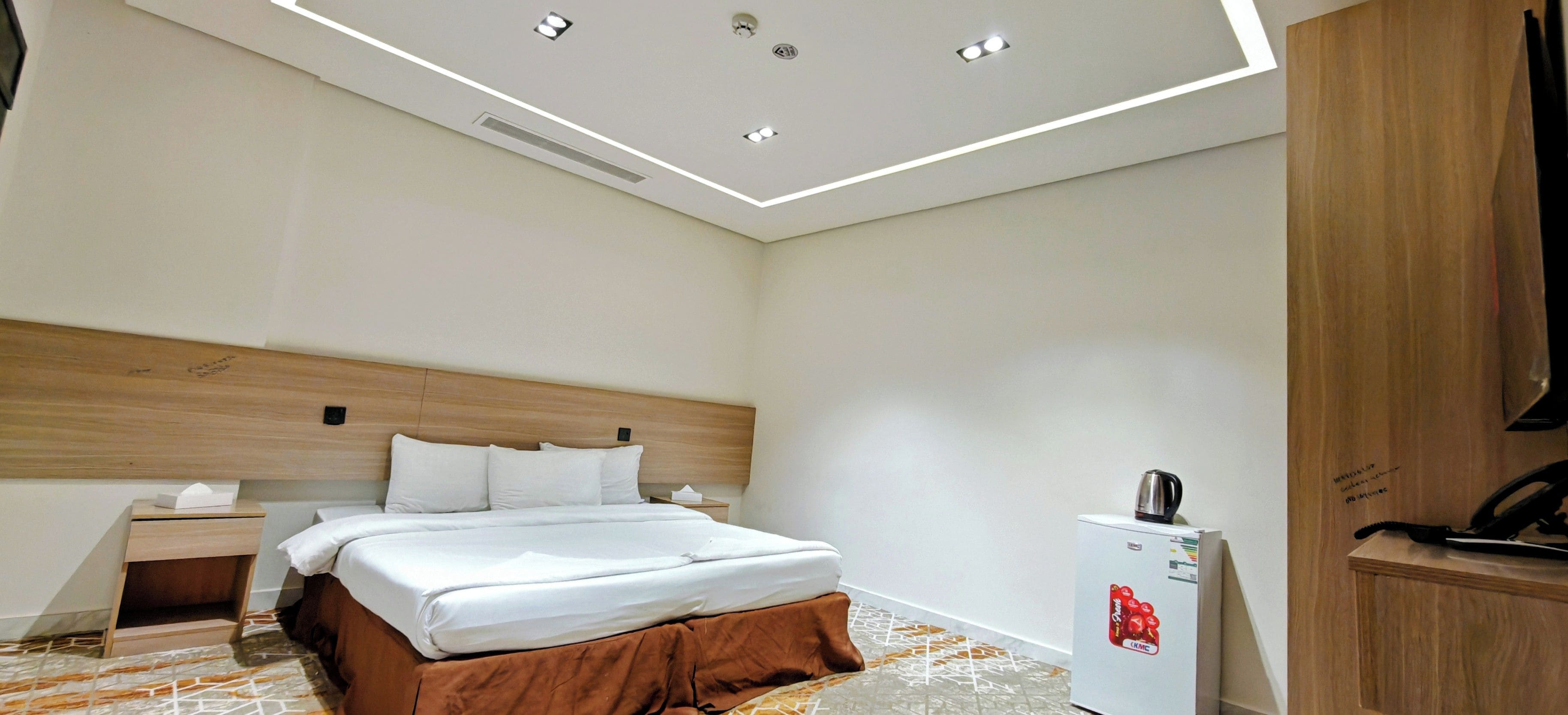 Een hotelkamer met een bed, nachtkastje, bureau, KMC-koelkast en een houten garderobe.