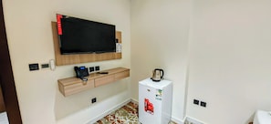 A hotel room with a flat-screen TV mounted on the wall, a wooden console table, a small refrigerator, and a tiled floor.