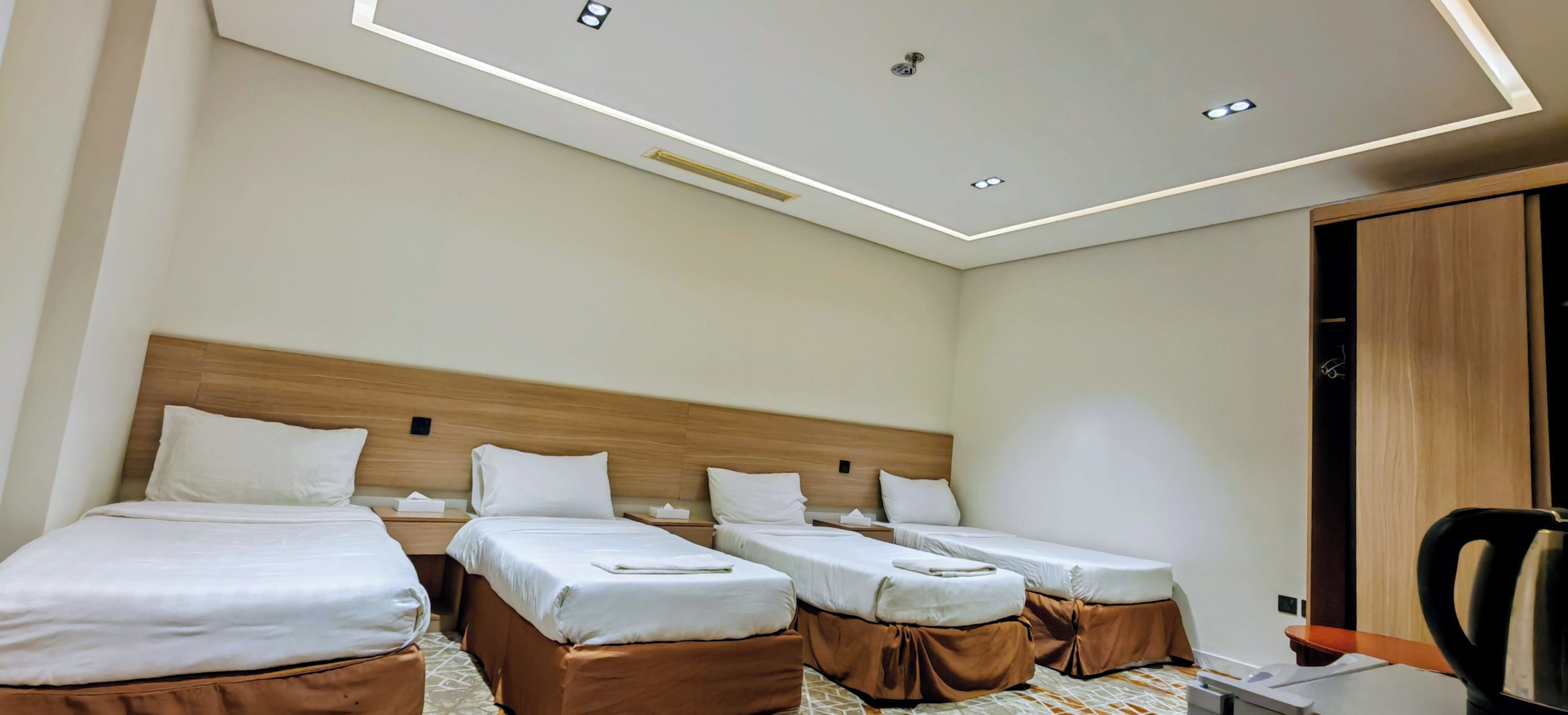 A hotel room with three single beds, a wooden headboard, a desk with a chair, and a ceiling with recessed lighting.
