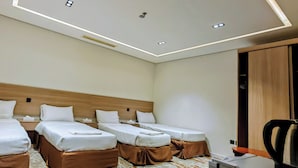 Habitación de hotel con tres camas individuales, cabecera de madera, escritorio con silla e iluminación empotrada en el techo.