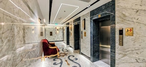 A marble-floored hallway with a red velvet chair, elevators, and emergency service signs.