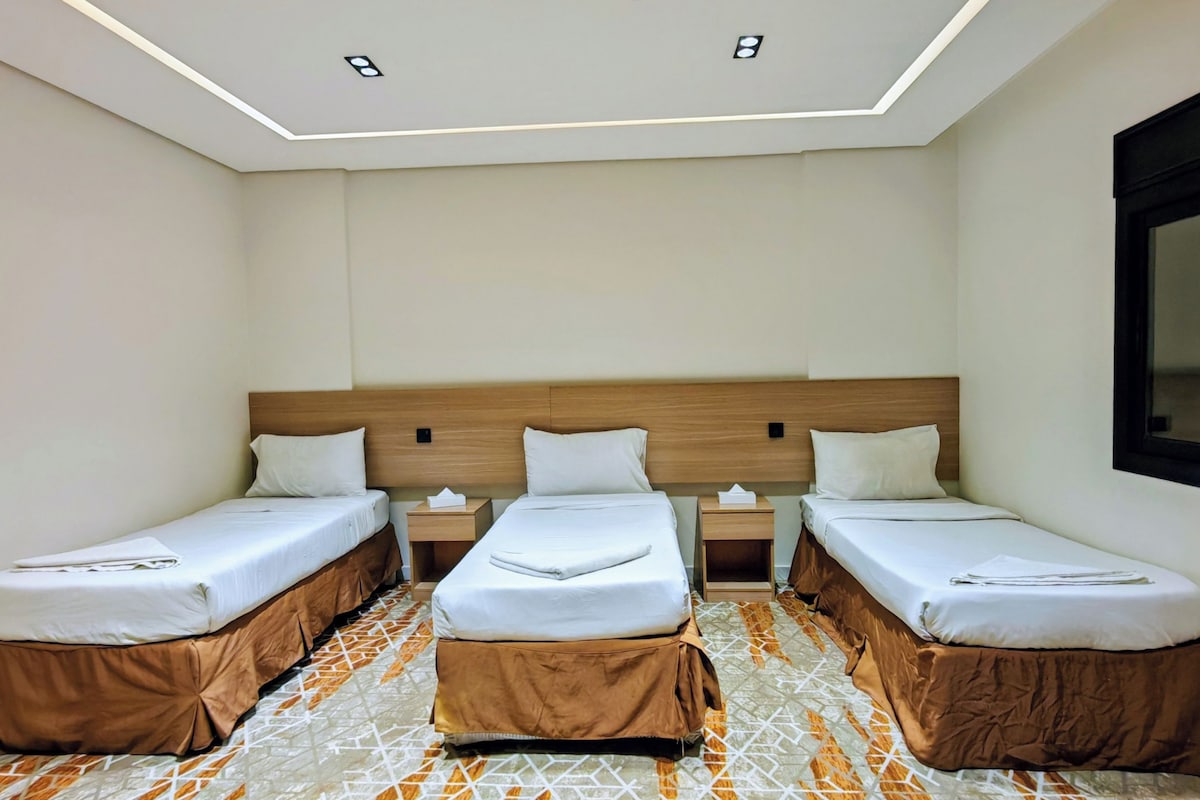 Ein Hotelzimmer mit drei Einzelbetten, einem Holz-Kopfteil, einem Fenster und einem gemusterten Boden.