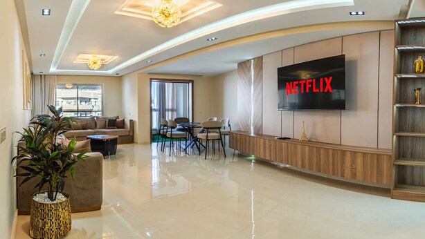 Ein modernes Wohnzimmer mit einem Flachbildfernseher, der 'NETFLIX' zeigt, einem Sofa, einem Couchtisch, einem Essbereich mit Stühlen und einer Zierpflanze.