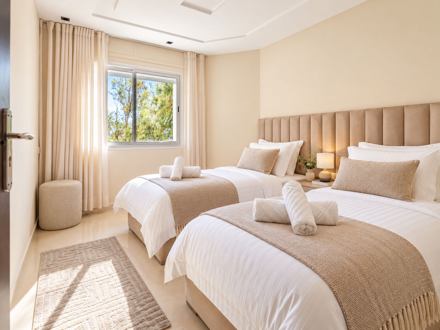 Een hotelkamer met twee bedden, een groot raam, een nachtkastje met lamp en een klein, rond voetenbankje.