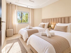 Une chambre d’hôtel avec deux lits, une grande fenêtre, une table de chevet avec une lampe et un petit pouf rond.