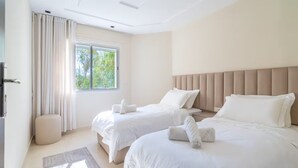 Ein Hotelzimmer mit zwei Betten, einem Fenster mit Vorhängen, einem Nachttisch und einem kleinen Hocker.