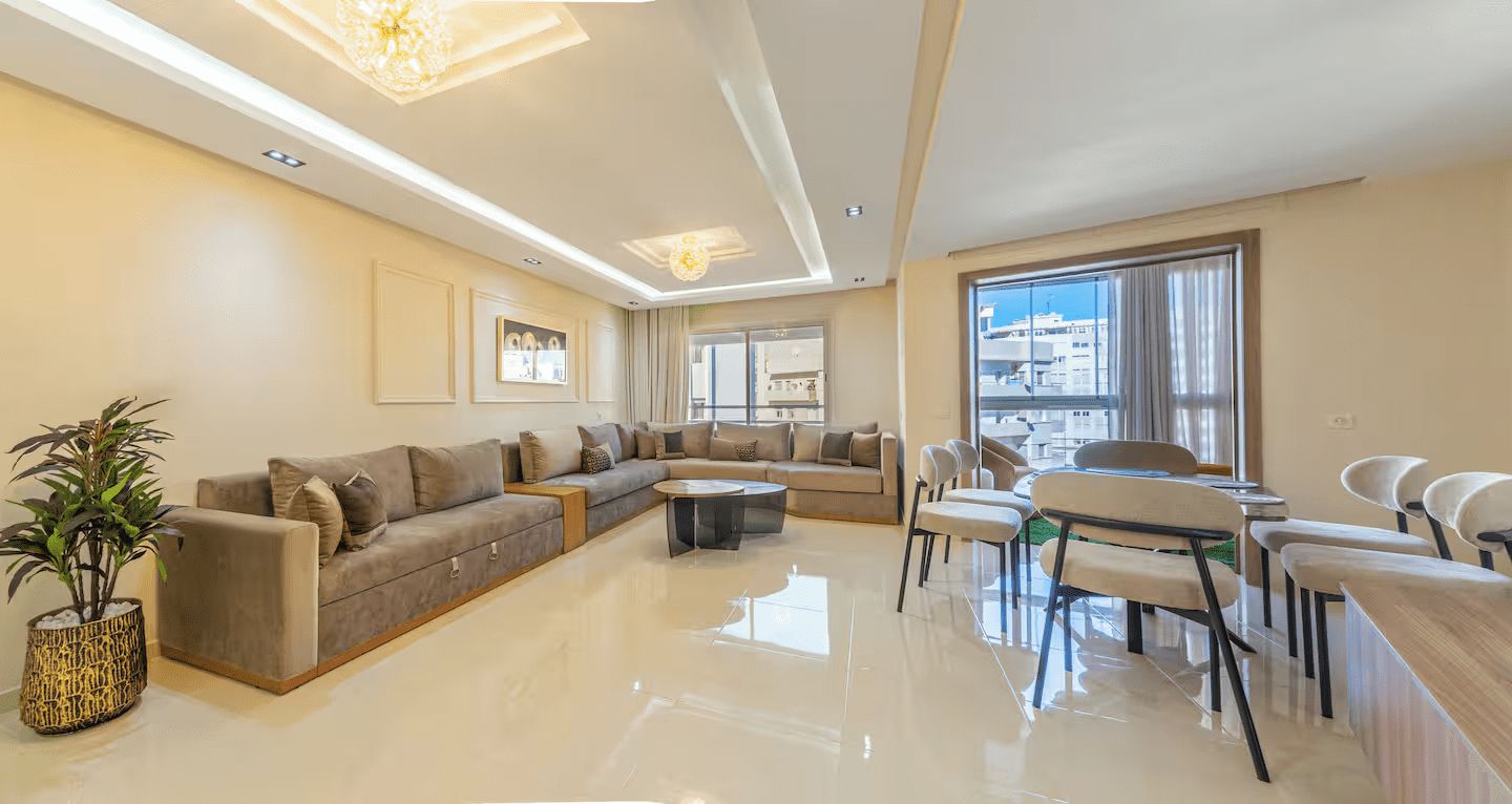 A modern living room with a sectional sofa, a dining area with chairs, and a large window providing a view of the city.