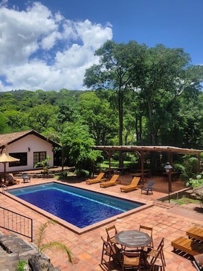 A pool area with a clear blue pool, wooden lounge chairs, a pergola, and a small building with a thatched roof.