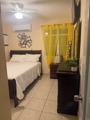 Ein Schlafzimmer mit einem Bett, einem Deckenventilator, einer Klimaanlage, einem Nachttisch mit Pflanze und einem Fenster mit gelben Vorhängen.