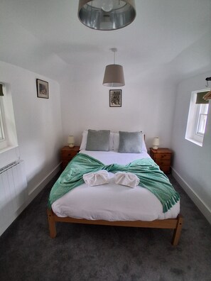 A bedroom with a bed, two bedside tables, a hanging lamp, a radiator, and two framed pictures on the wall. - Cottage Two (Dover)