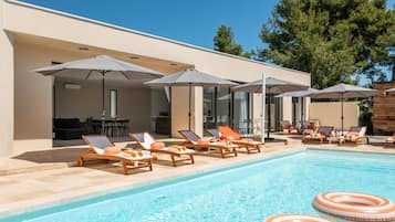A poolside area with lounge chairs and umbrellas, a clear blue pool, and a modern building facade.