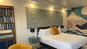 Quarto moderno com cama grande, mesa de cabeceira com luminária, estante e um mural decorativo na parede.