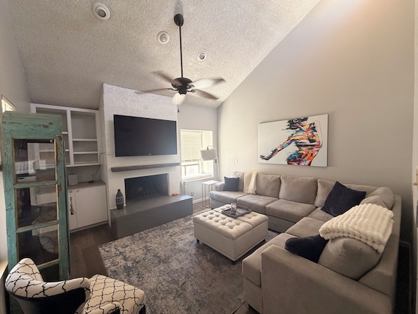 A living room with a fireplace, a television, a ceiling fan, and a sofa. - Vaulted ceilings, brand new appliances, overlooking pool/hot tub of complex. (Scottsdale)