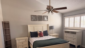 Ein Schlafzimmer mit einem Bett, Nachttischen, einer Bank, einem Deckenventilator und einem Fenster mit Jalousien.