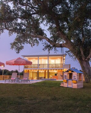 Una casa moderna con un gran árbol en primer plano, una piscina y mobiliario de exterior.