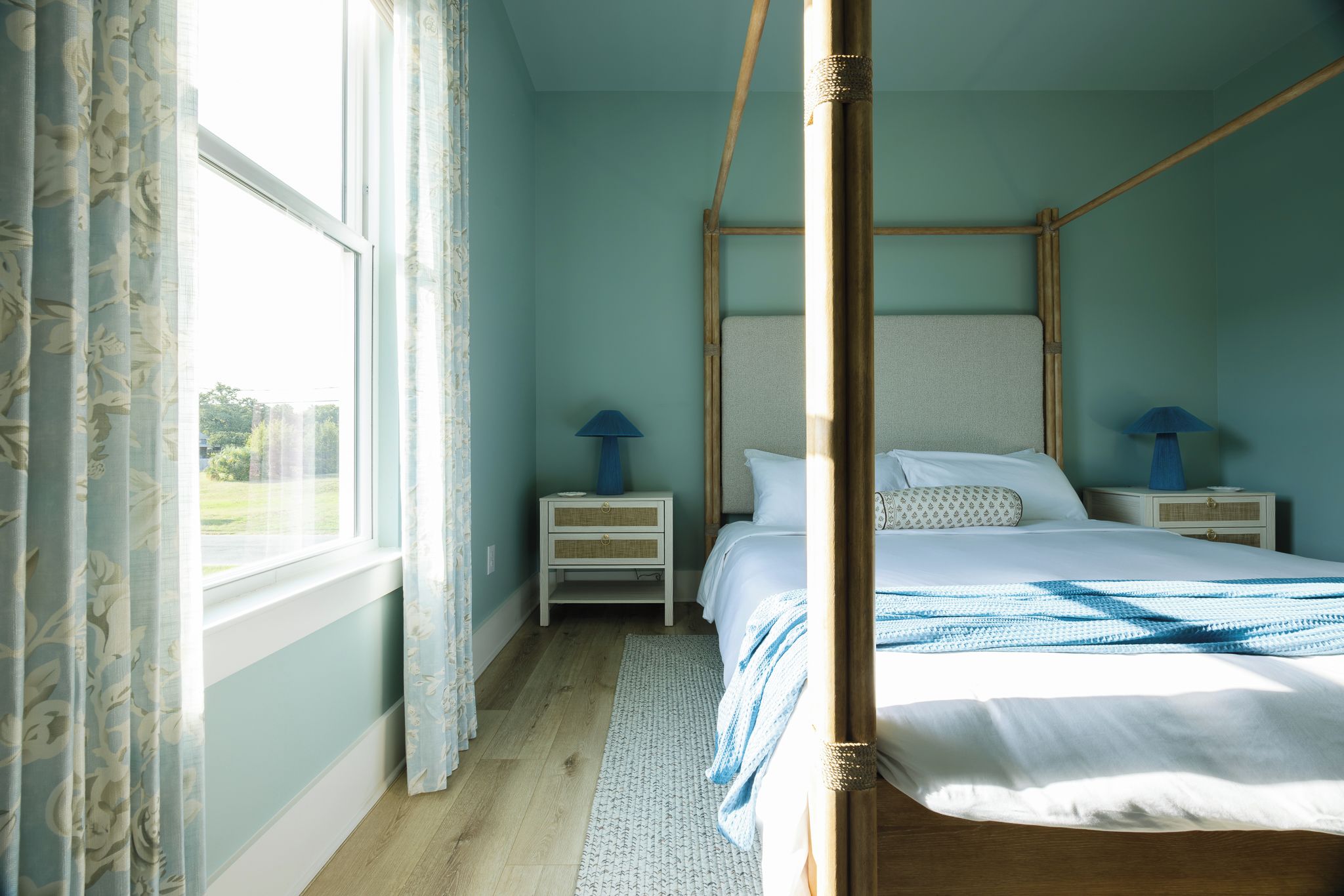 A four-poster bed with white linens, two bedside tables with lamps, a window with curtains, and a view of greenery outside.