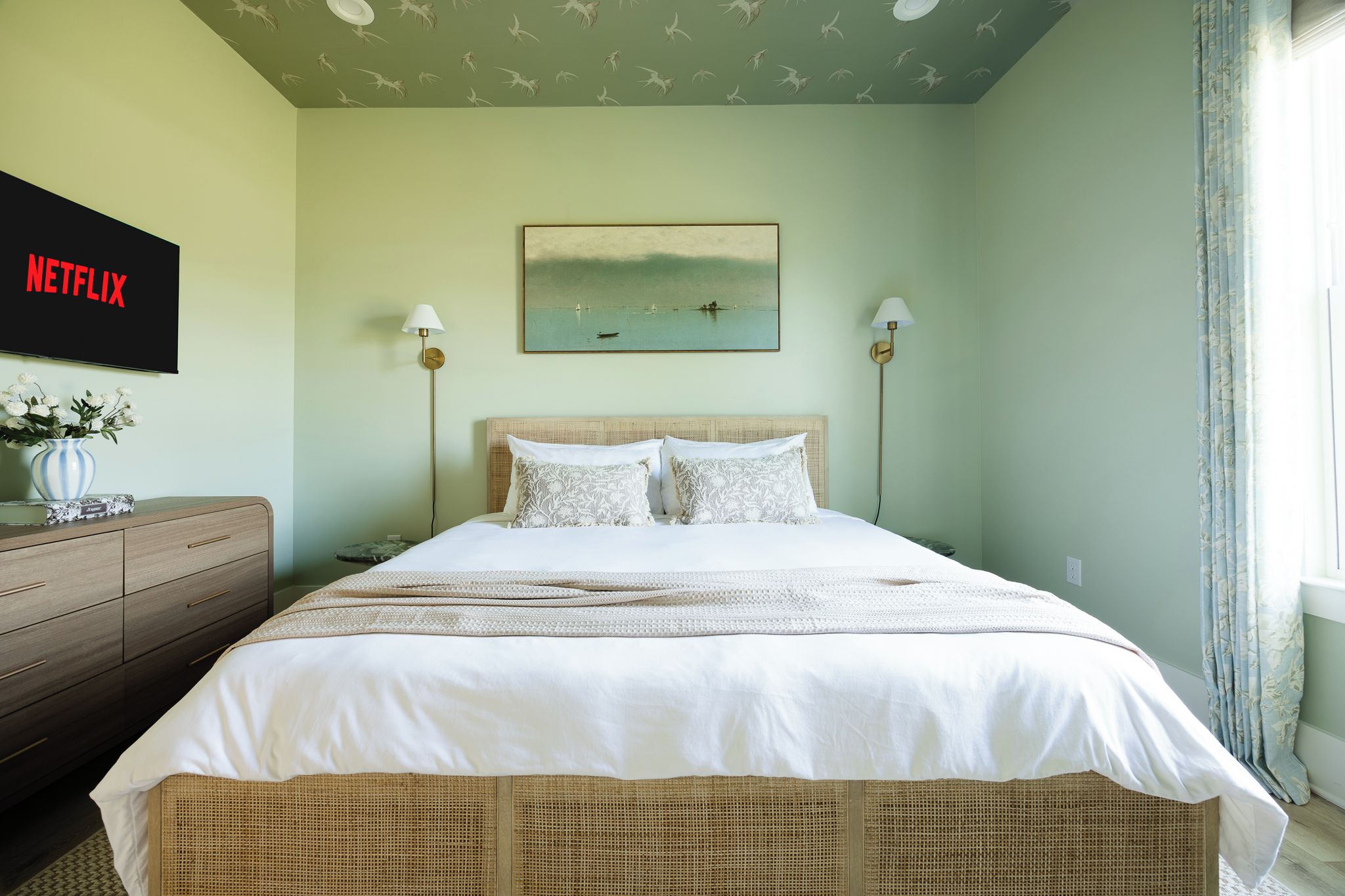 Un dormitorio con cama, televisión montada en la pared, un cuadro y una ventana con cortinas.