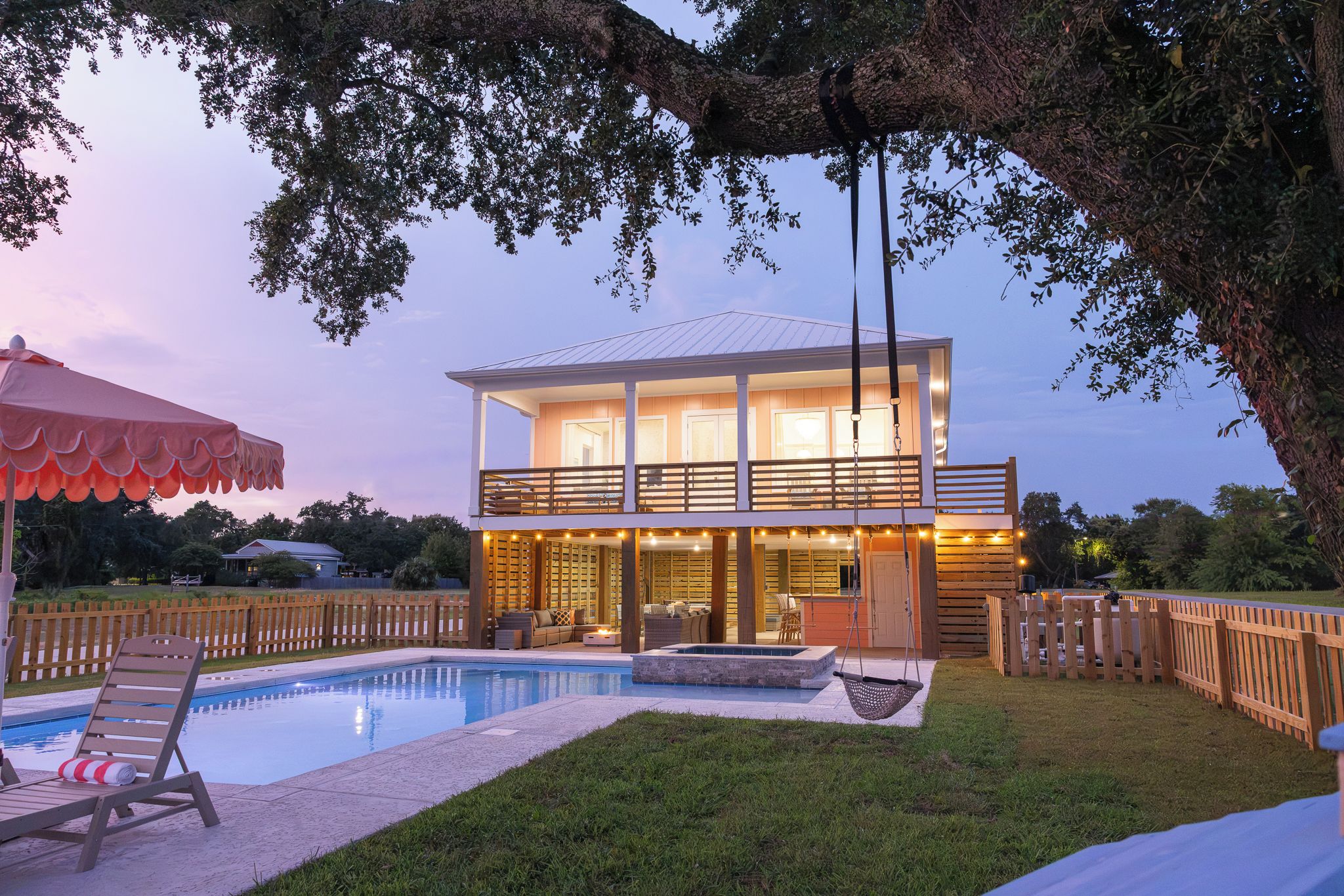 A two-story house with a swimming pool, outdoor seating, and a large tree.