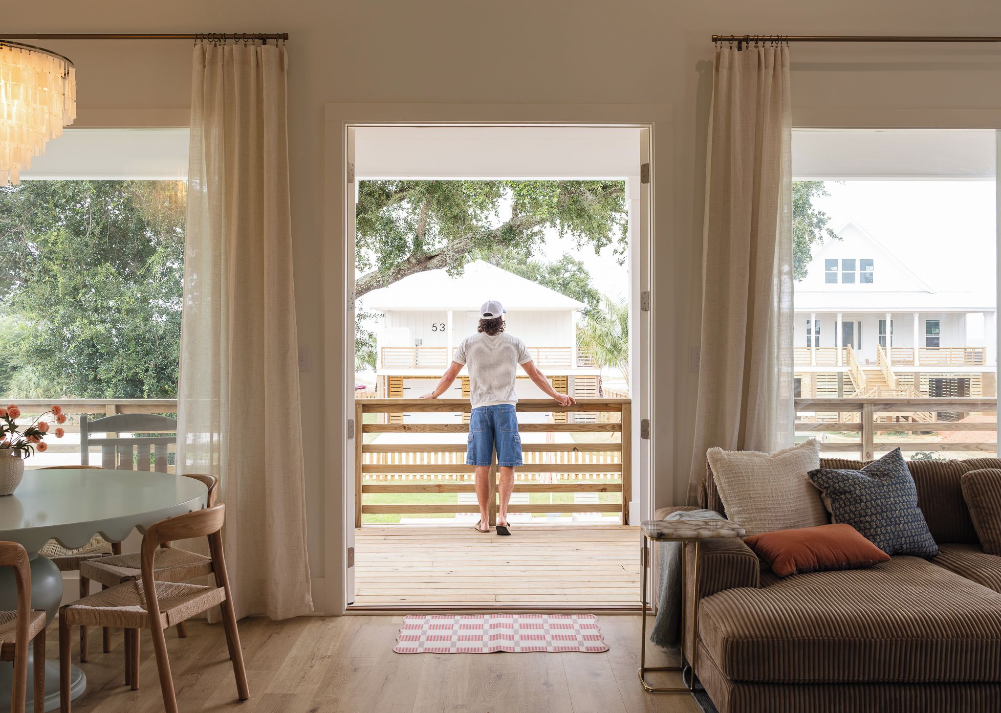 A room with a wooden deck, a dining area with wicker chairs, a sofa with cushions, and a large window with curtains.