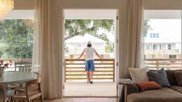 A room with a wooden deck, a dining area with wicker chairs, a sofa with cushions, and a large window with curtains.
