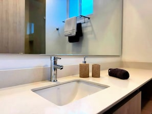 A modern bathroom with a white sink, a chrome faucet, and a mirror above it. There are two cylindrical containers on the countertop, and a rolled-up towel is placed on the sink's edge. - Deluxe Suite with Jacuzzi - Pozos Colorados (Magdalena)