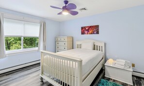 A bedroom with a crib, a dresser, a nightstand, a ceiling fan, and a window with blinds. - Quechee Art and Trail Retreat (Quechee)
