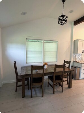 A dining area with a wooden table and chairs, two windows with blinds, a hanging light fixture, and a kitchen area with cabinets.